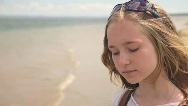 Close up portrait of beautiful young woman teenager. Hair blowing fluttering in wind on beach in slow motion. She is attractive and calm, sad, thoughtful. She has sunglasses.