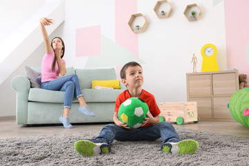 Little boy playing with ball at home while his nanny is taking selfie