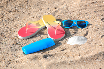 Fototapeta na wymiar Sunglasses, bottle with sunscreen cream and flip flops on sand. Beach object