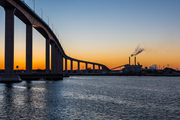 Fototapeta premium The Jordan Bridge over the Elizabeth River in Virginia is silhouetted against a vibrant orange sunset, industry and factories in the background