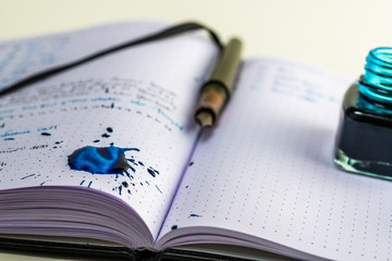 Notebook with writing and a fountain pen with blue ink, ink splattered on the paper making a mess and a large blue ink blot