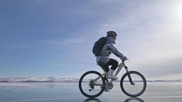 Woman is riding bicycle on the ice. The girl is dressed in a silvery down jacket, cycling backpack and helmet. Ice of the frozen Lake Baikal. The tires on the bicycle are covered with special spikes