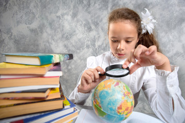 The schoolgirl in school uniform looking at a globe through a magnifying glass and writing in a notebook.