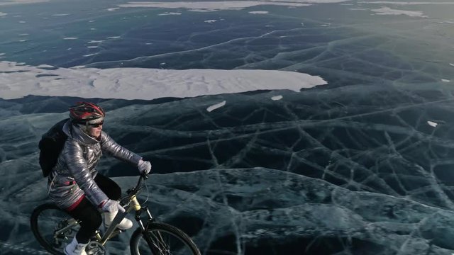 Woman is riding bicycle on the ice. The girl is dressed in a silvery down jacket, cycling backpack and helmet. Shooting with a quadrocopter drone. The tires on the bicycle are covered with special