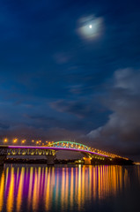 Auckland harbour bridge at night lit up in bright colours, with the moon.