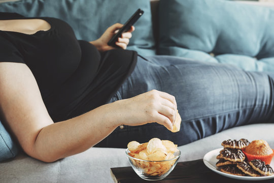 g, sedentary lifestyle, compulsive overeating. Obese woman laying on sofa with smartphone eating chips