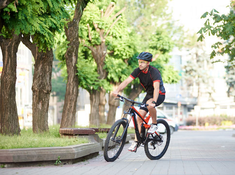 Athletic guy bicyclist in cycling clothes and protective gear riding on bicycle along empty city streets, green trees around. Sportsman performing morning outdoor exercise and training.