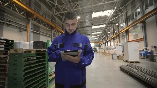 A man is working in a factory with a digital tablet, he is dressed in a blue uniform. factory worker.