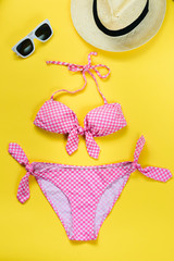 Top view of two pieces pink chequered swimming suit, white sunglasses and straw hat over yellow...