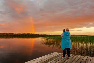 Summer evening. A woman stands on the dock and takes a photo of the rainbow.