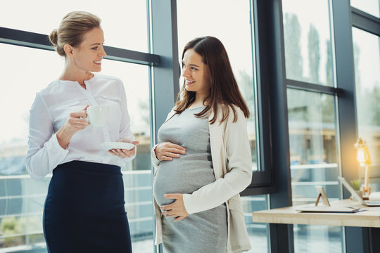 Sharing plans. Pretty cute pregnant woman standing with her hand on a belly while telling her curious boss about plans for the future
