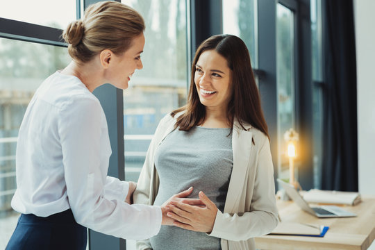 Touching Belly. Cheerful Kind Friendly Woman Smiling While Being At Work And Touching The Belly Of Her Pregnant Colleague