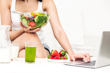 Fototapeta na wymiar Woman holding healthy fresh salad in a bowl and working on a laptop computer while relaxing on sofa at home.dieting concept.healthy lifestyle with green food