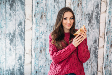 A sweet, cheerful European-looking girl dressed in a stylish warm jacket is going to eat her sandwich with contentment, on a wooden background.