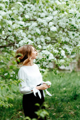Cute spring portrait. Young business woman in a business office suit on the background of Apple trees in bloom. Girl holding a white flower.