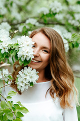 Gentle spring portrait of a young blonde woman in a white shirt on a background of blooming apple...