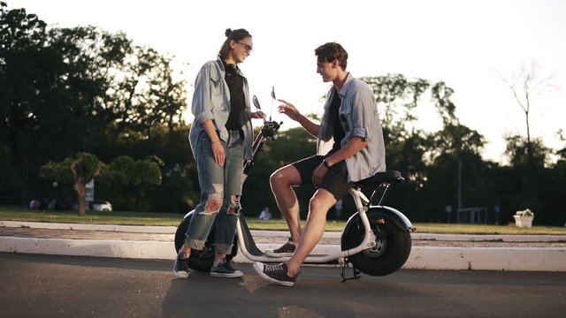 Two young stlylish friends are hanging out together. The boy is sitting on bike and talking to a beaitiful blonde girl in front. Happy, carefree, smiling