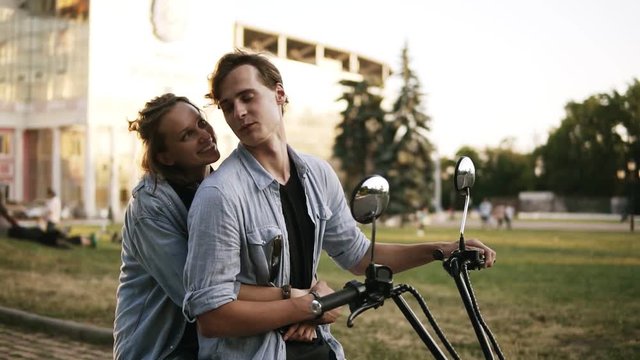 Overview footage of a beatuful couple sitting on a bike. Girl is hugging man from the back. Wearing black T shirt and blue shirt. Summer day