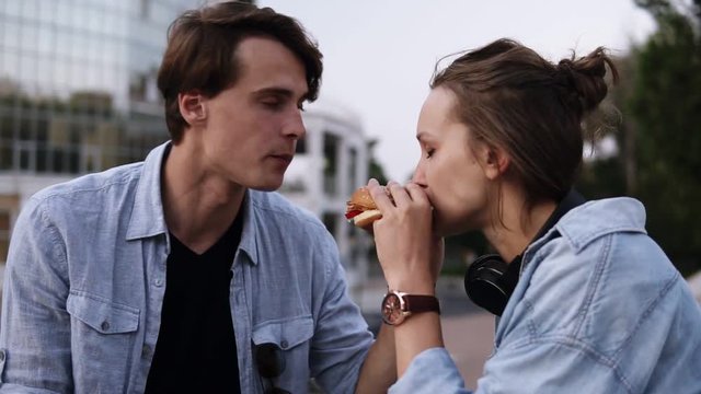Beautiful young couple enjoying a meal together. Eating hamburger together from the sides. Evening dusk, park side
