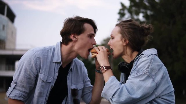 Young people are biting a big burger together. Sharing a food. Evening dusk in the park. Couple, dating, spending time together