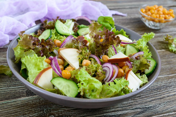 Spicy healthy salad of fresh greens, red onion, chickpeas, smoked cheese (sausage cheese), cucumbers in a bowl on a wooden background. Vegetarian dish. Close-up.