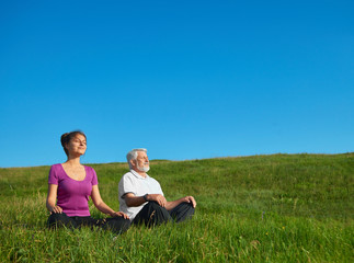 Young girl and old man meditating sitting in the field. Sitting in lotus pose on bright saturated sky background. Looking positive, satisfied, feeling good, concentrated. Calm and silent.