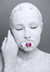 a large portrait of a girl covered with white powder with a flower of orchids in her mouth on a gray background. Art photo of a young woman who looks like a sculpture, a white statue