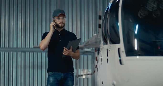 Male mechanic in uniform examining helicopter. Talking on the phone during preflight inspection at the airport. 4K UHD