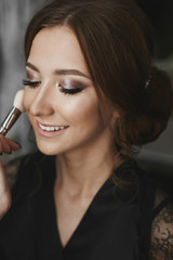 Makeup artist applying makeup on face of a beautiful and cheerful brunette model girl with charming smile and closed eyes, in black peignoir sitting in interior