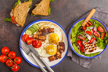 Vegetable salad with goat cheese, sandwiches with cheese and vegetables and fried eggs with bacon. Beautiful bright breakfast on a dark background
