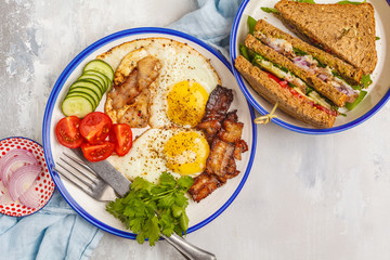 Fried eggs with bacon and a sandwich with meat, cheese and vegetables on white background, top view.