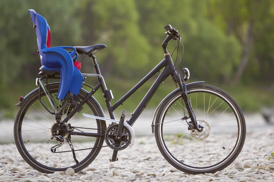 Fototapeta Close-up of new modern black bicycle with blue plastic child seat standing on side stand on lit by sun pebbles on blurred green trees summer bokeh background. Tourism and family traveling concept.