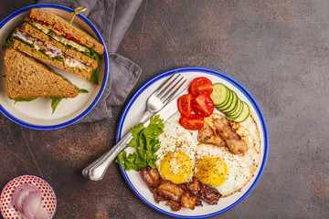 Fried eggs with bacon and a sandwich with meat, cheese and vegetables on dark background. Delicious hearty breakfast.