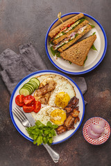 Fried eggs with bacon and a sandwich with meat, cheese and vegetables on dark background. Delicious hearty breakfast.