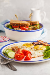Fried eggs with bacon and a sandwich with meat, cheese and vegetables on white background. Delicious hearty breakfast.