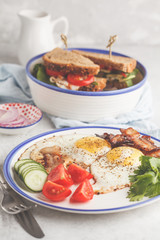 Fried eggs with bacon and a sandwich with meat, cheese and vegetables on white background. Delicious hearty breakfast.