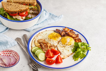 Fried eggs with bacon and a sandwich with meat, cheese and vegetables on white background. Delicious hearty breakfast.