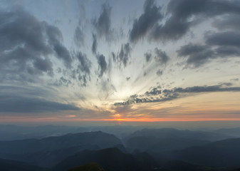 Fantastic sunrise or sunset over green mountain ridge covered with dense blue fog. Bright orange sun raising in  soft cloudy sky over distant horizon. Beauty of nature, tourism and traveling concept.