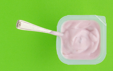 Yogurt in plastic cup close up with small silver spoon, top view photo of strawberry yoghurt on green background