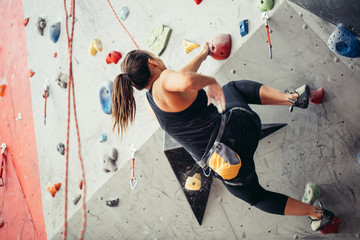 Unrecognizable athlete climber woman exercising on grey practical wall with colourful hooks, indoor low angle shot, bouldering, rear view.