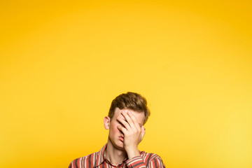 facepalm. ashamed abashed man covering his face with hand. portrait of a young guy on yellow background popping up or peeking out from the bottom. copy space for advertising. - obrazy, fototapety, plakaty