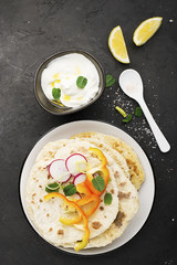Traditional homemade flat breads for a snack with fresh seasonal vegetables, herbs and yoghurt sauce with olive oil and rose salt on the background. Top view