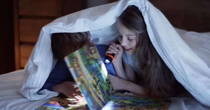 Close up of the cute small sister and brother with a flashlight looking at the colourful book with pictures and flipping pages under the blanket at night. Inside.