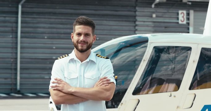 Portrait of commercial pilot in uniform standing near small private helicopter on a landing point. 4K UHD
