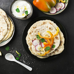 Traditional homemade flat breads for a snack with fresh seasonal vegetables, herbs and yoghurt sauce with olive oil and rose salt on the background. Top view.
