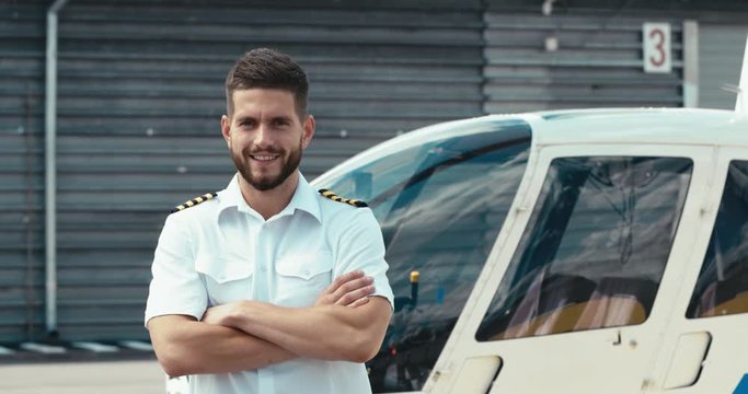 Portrait of commercial pilot in uniform standing near small private helicopter on a landing point. 4K UHD