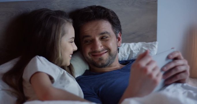 Close up of the cheerful father and his small daughter lying in the bed before falling asleep and smiling while watching funny video on the smartphone. Indoors.