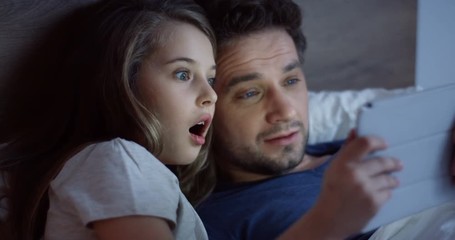 Close up of the small cute girl lying in the bed before sleeping with her handsome father and watching scary video on the tablet device. Inside.
