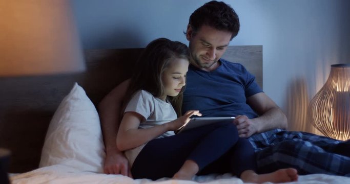 Caucasian father lying on the bed with his cute little daughter in the evening and girl scrolling and taping on the tablet device. Indoors.
