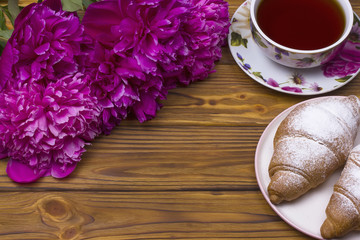 Purple peony flowers with croissant and teacup on brown wooden background.